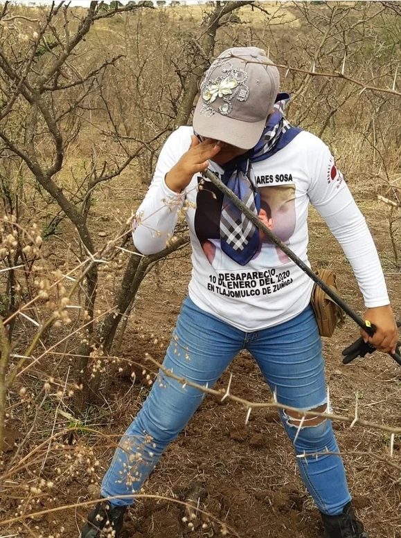 A searching mother with a probe in the scrubland, wearing a shirt with the photo of her disappeared son